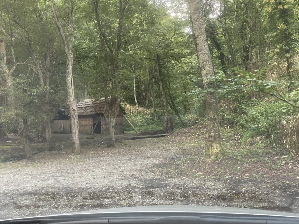 View of the parking area and restored houses at the Sankebetsu Bear Attack Site, surrounded by lush forest.