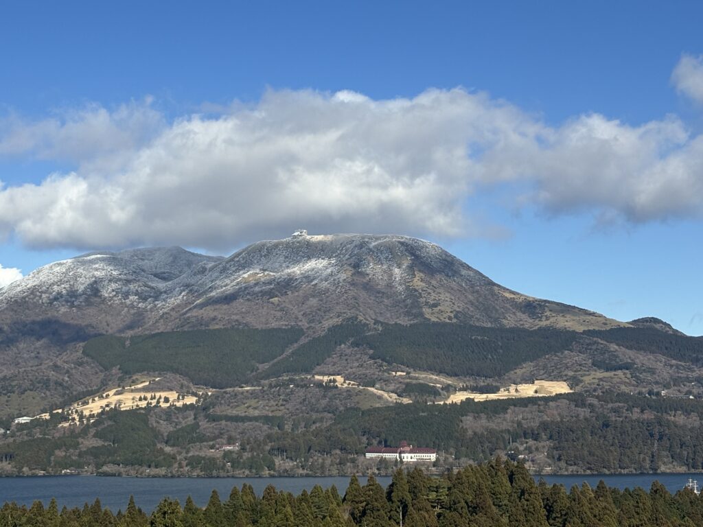 Snow-capped mountain overlooking Lake Ashi in Hakone, with forested slopes and a building visible near the shoreline