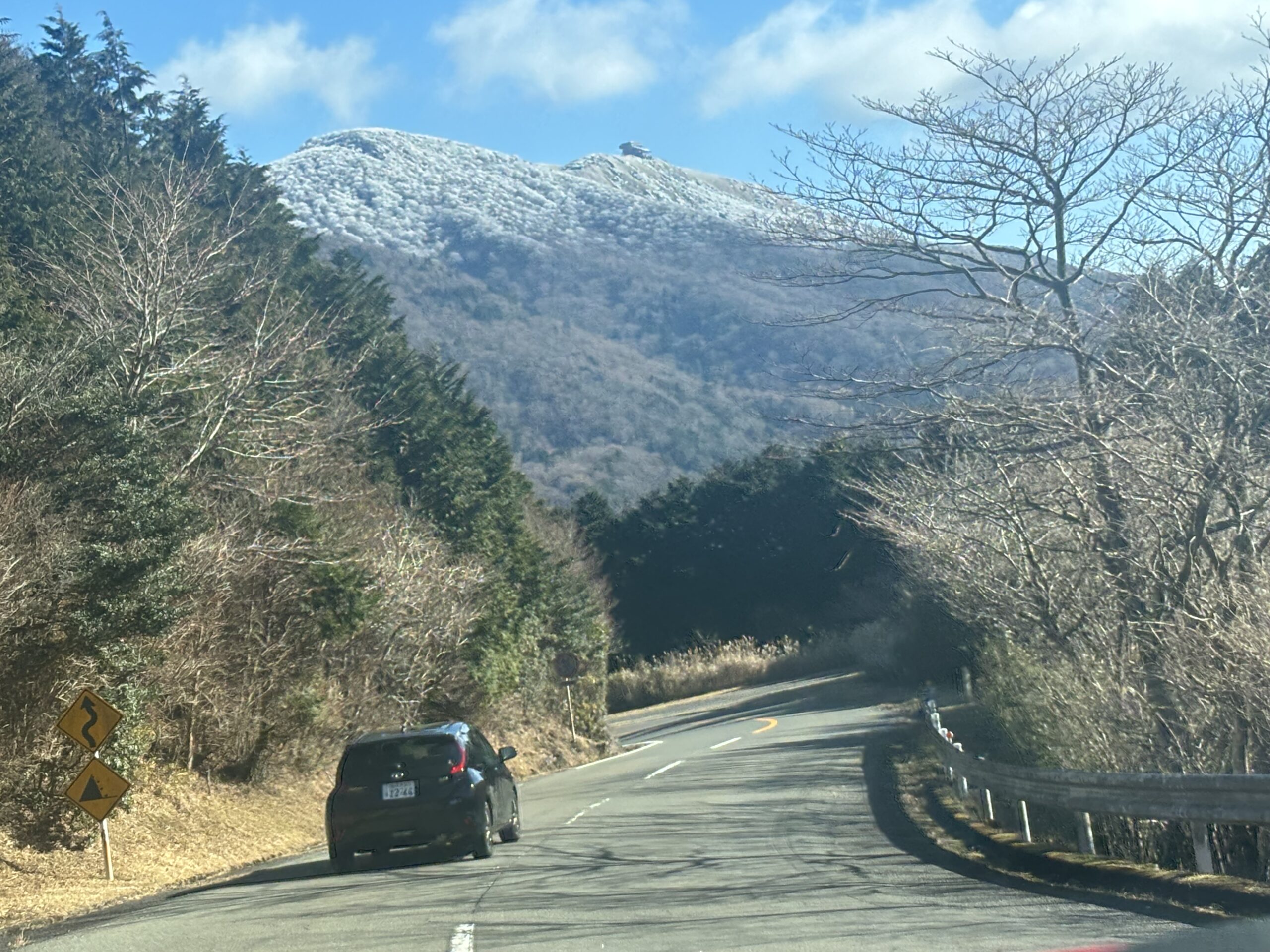 Winding mountain road with guardrails leading uphill, snow visible on the mountain peak, and a car driving ahead