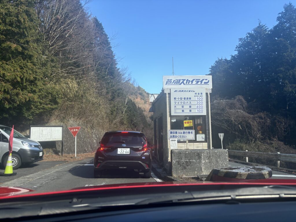 "Toll booth entrance with pricing sign in Japanese showing fees for different vehicle types, and a car waiting at the gate