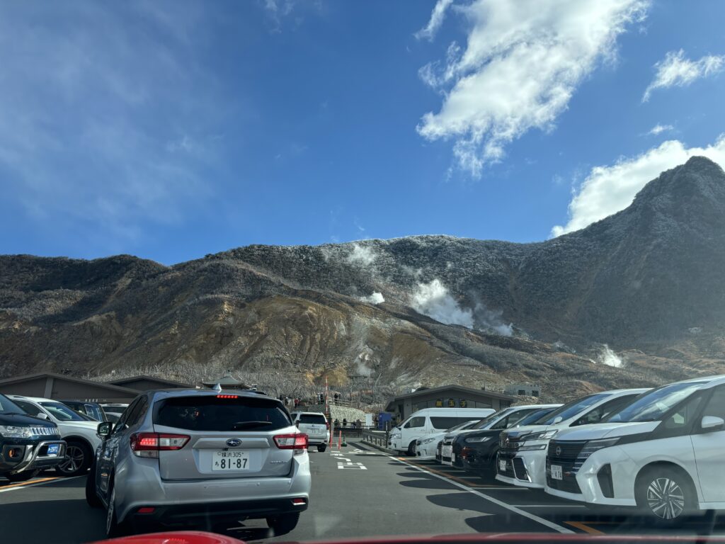 "Crowded parking lot with steam rising from volcanic vents on mountainside in background, filled with various cars and vans